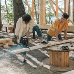 Two adult men working on wood construction project outdoors, using a chainsaw. — examplearticle