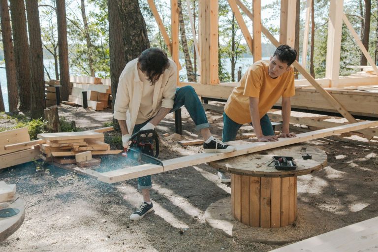 Two adult men working on wood construction project outdoors, using a chainsaw. — examplearticle