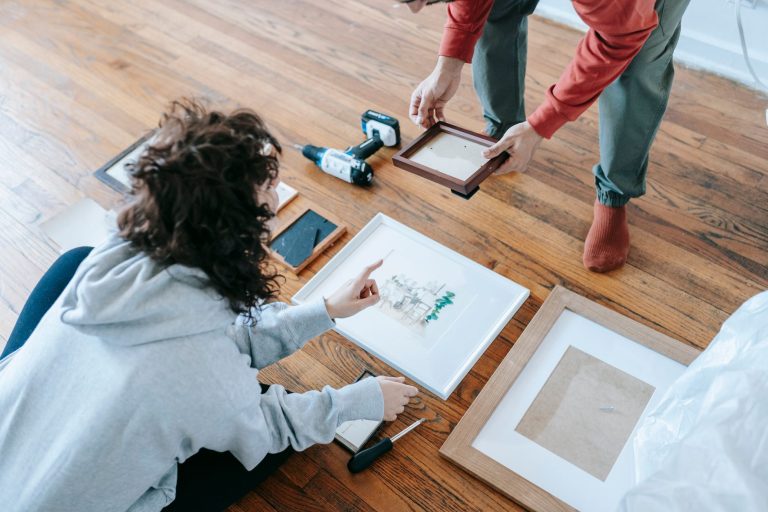 A couple organizing picture frames on a wooden floor, preparing for interior decoration or renovation. — Recenzije i savjeti